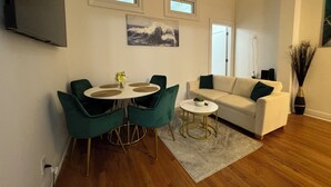 A modern living room with a round dining table, green chairs, a white sofa, and a small coffee table. There are two windows and a painting on the wall.