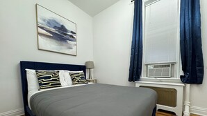 A neatly made bed with a grey bedspread and decorative pillows, a bedside table with a lamp, a window with blue curtains, and a wall-mounted air conditioning unit.