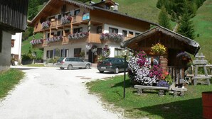A traditional chalet-style building with balconies adorned with flowers, a gravel driveway, and a mountainous backdrop.