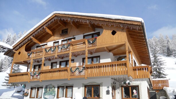 A multi-story wooden chalet with balconies, a stone entrance, and snow-covered surroundings.