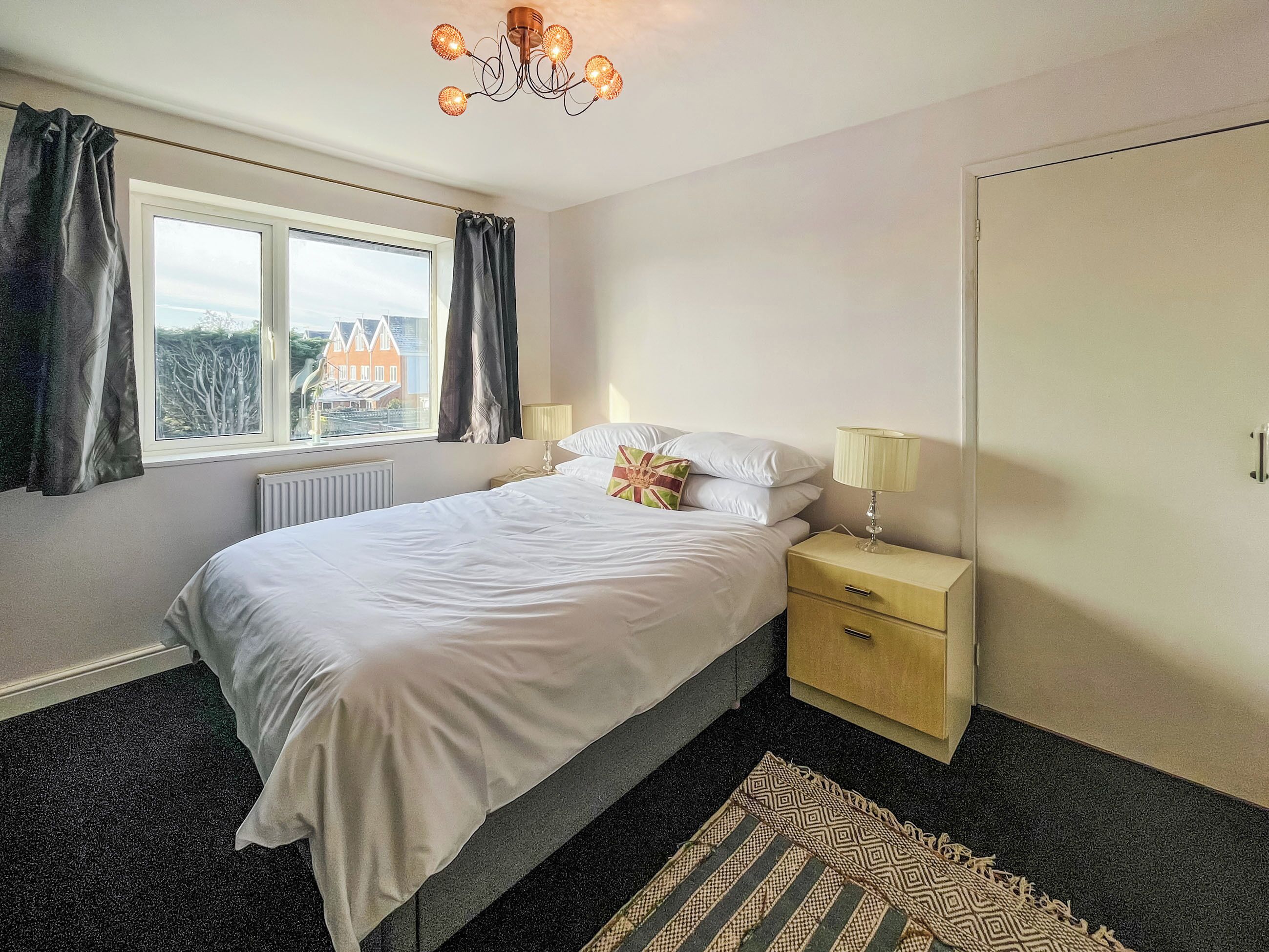 A bedroom with a bed, bedside tables, a window with curtains, a radiator, and a patterned rug.