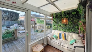 Un espace de détente intérieur, chaleureux, avec un canapé confortable, un petit tabouret en bois et une porte vitrée donnant sur une terrasse extérieure.