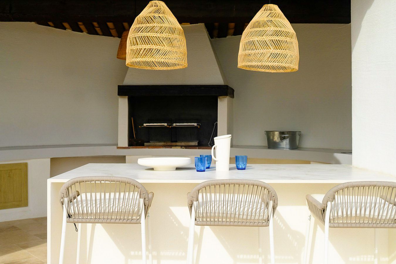 An outdoor kitchen with a built-in grill, white bar stools, and hanging wicker pendant lights.