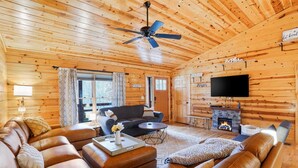 A cozy log cabin interior with a leather sofa, a coffee table, a fireplace, and a ceiling fan. - Honeybee Hideaway | Hot Tub, Game Room, Pond (Logan)
