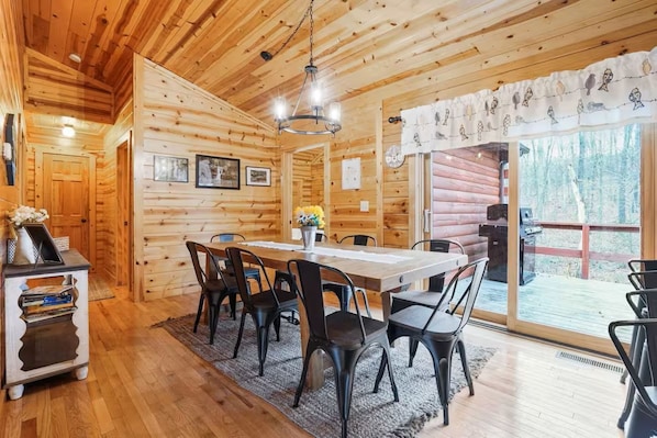 A wooden cabin interior with a dining table, chairs, a chandelier, and a view of the outdoors.