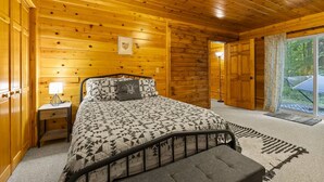 A wooden cabin bedroom with a bed, nightstand, and a view of the outdoors through a door. - Honeybee Hideaway | Hot Tub, Game Room, Pond (Logan)