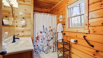 A wooden cabin bathroom with a sink, mirror, towel rack, shower curtain, and window.