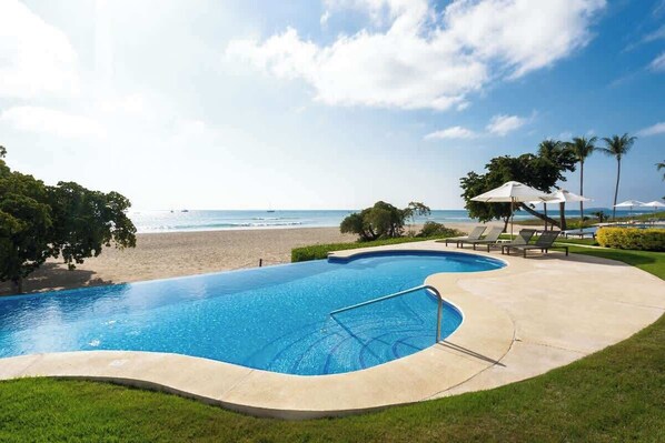 A swimming pool with a curved edge, a beach view, and lounge chairs.