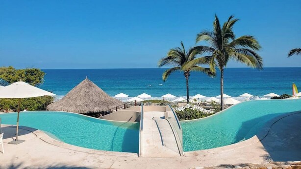 A resort pool area with a clear view of the ocean, palm trees, and thatched umbrellas.