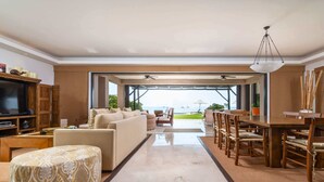 A spacious living room with a sofa, ottoman, and dining area with chairs. A television and entertainment unit are visible.