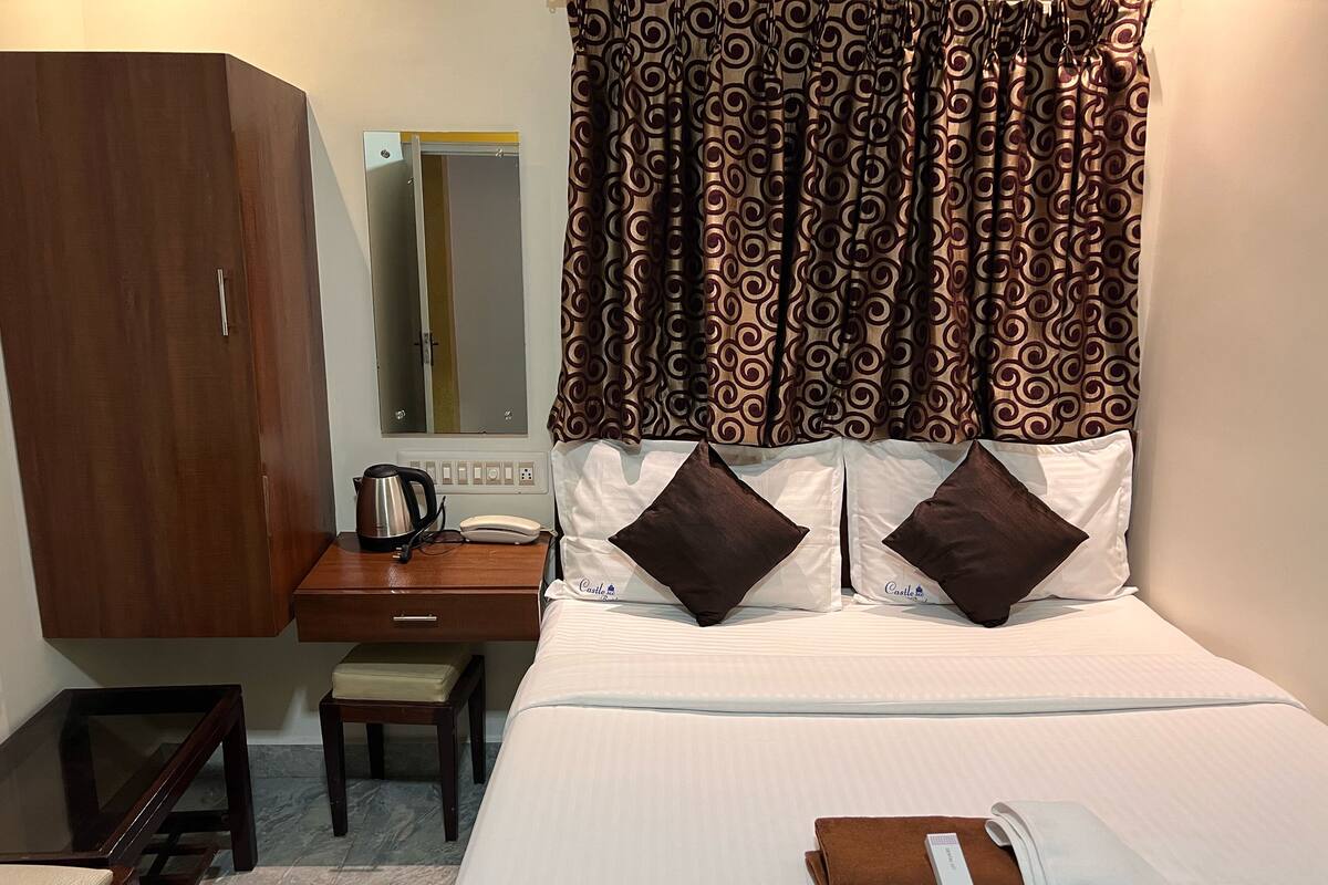 A hotel room with a bed, a desk with a kettle, a telephone, and a brown chair.