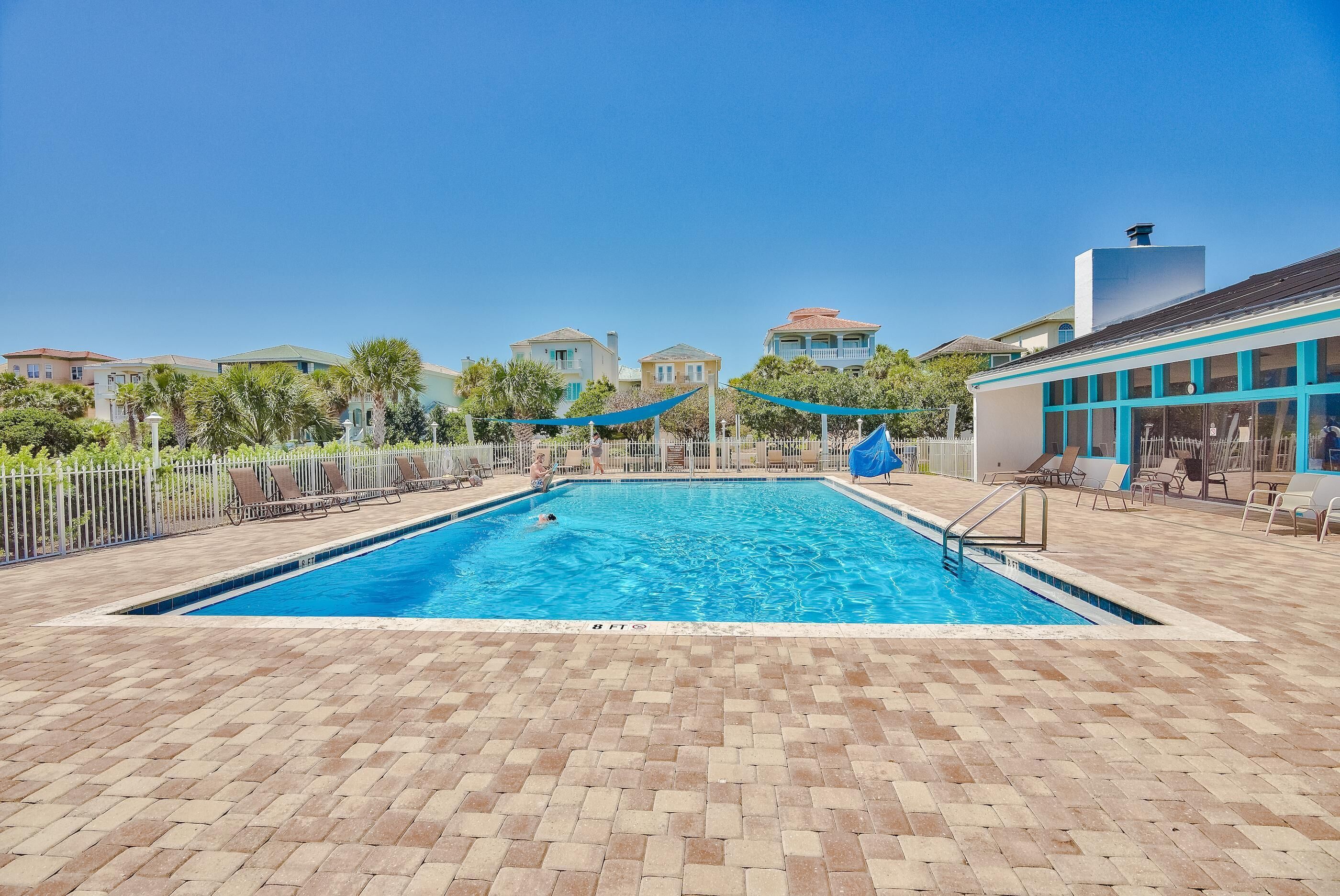 A rectangular swimming pool with a tiled deck, surrounded by a white fence and palm trees.