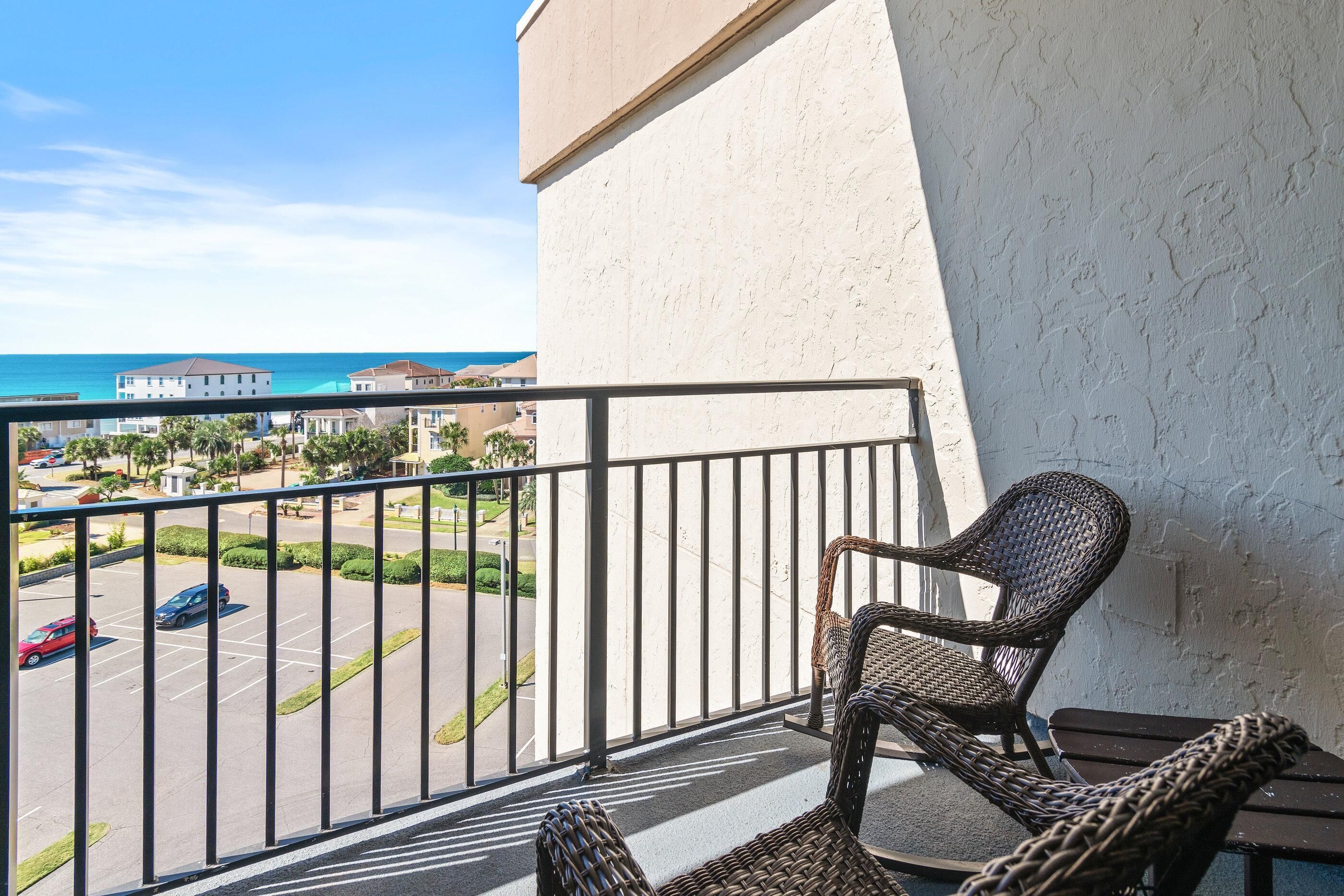 A balcony with wicker chairs, a view of a parking lot, and buildings in the background.