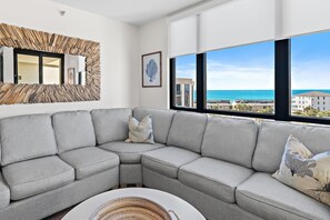 A modern living room with a grey sectional sofa, a woven coffee table, and a large window offering a view of the sea and buildings. - Enclave 703b- Sugar Sands (Destin)