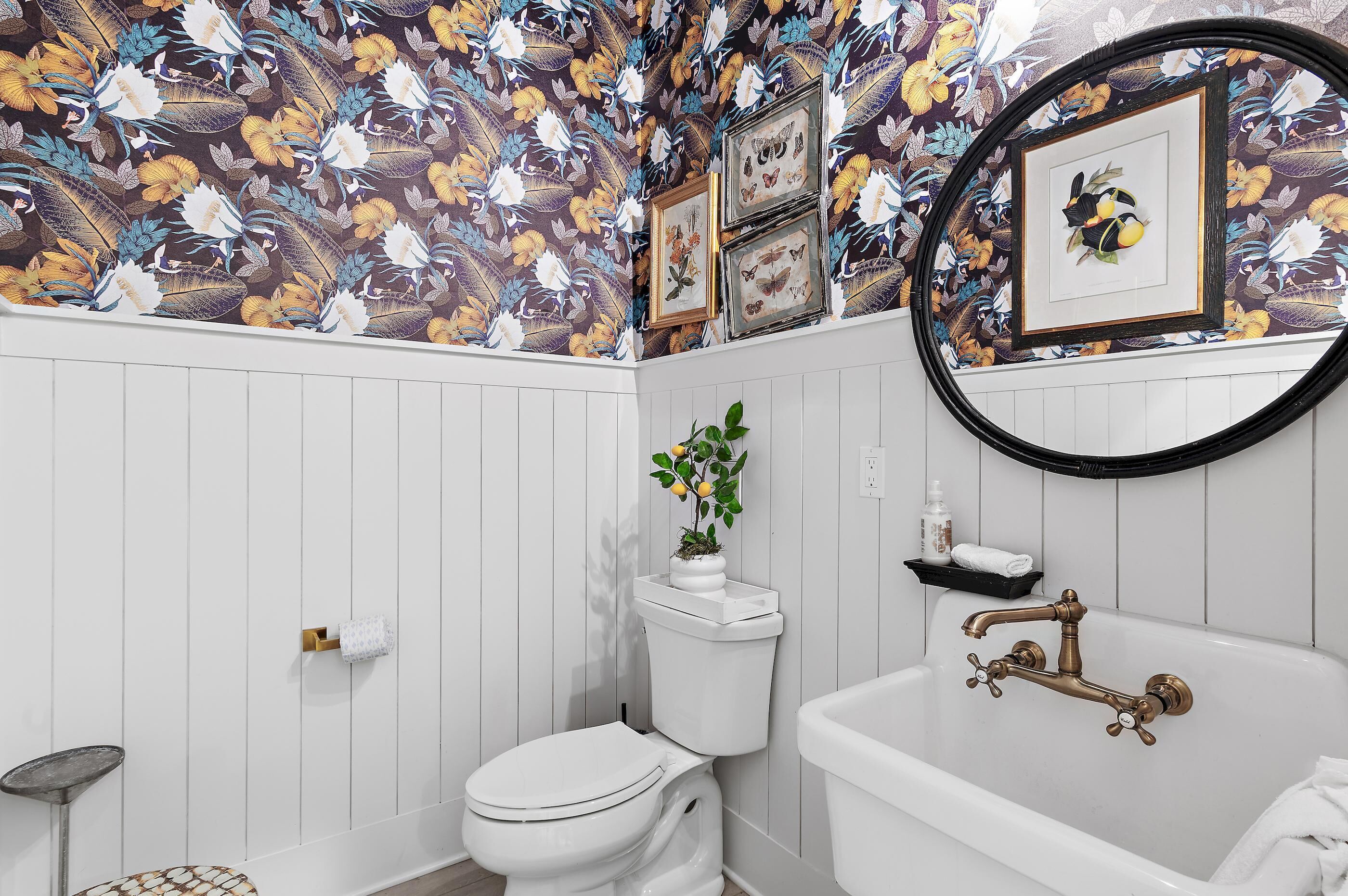 Une salle de bain comprenant des toilettes, un lavabo et une baignoire, avec un papier peint à motifs floraux et des tableaux encadrés accrochés au mur.