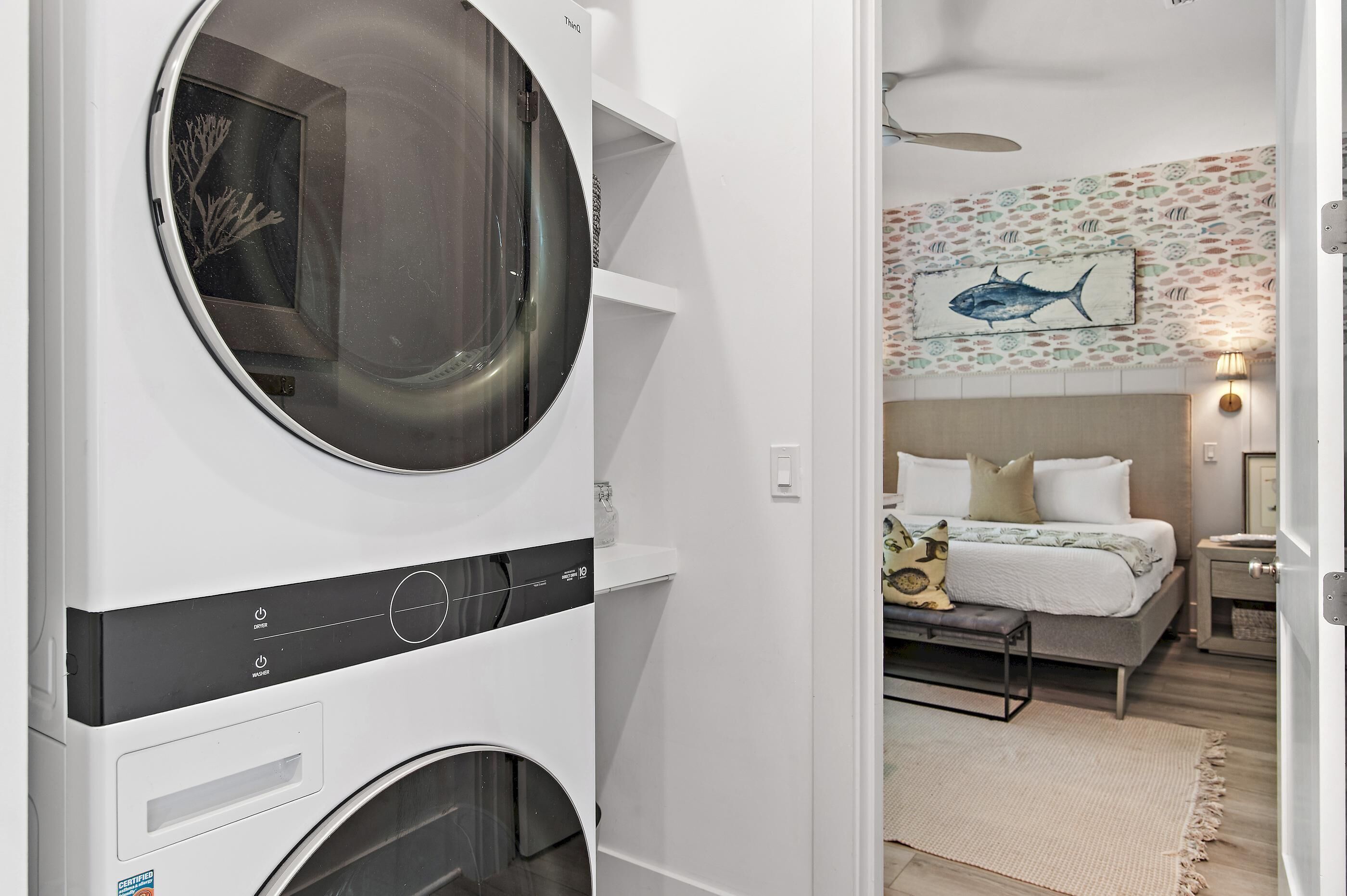 Een wasruimte met een voorlader wasmachine en droger, een bed met hoofdeinde, een nachtkastje en een muur met een vis-thema kunstwerk.