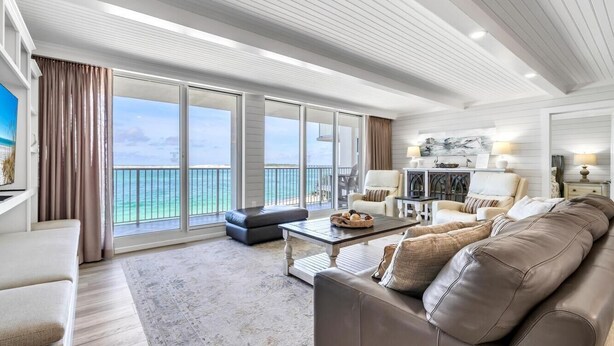 A spacious living room with a large window overlooking the sea, a leather sofa, a coffee table, and a dining area with a chandelier.