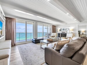 A spacious living room with a large window overlooking the sea, a leather sofa, a coffee table, and a dining area with a chandelier. - Waterview Towers 201 (Destin)