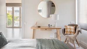 Un dormitorio con cama, escritorio, silla, espejo redondo y ventana con vista a edificios.