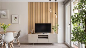 Una sala moderna con un televisor en una pared revestida de madera, una zona de comedor con mesa y sillas, y un ventanal con vistas.