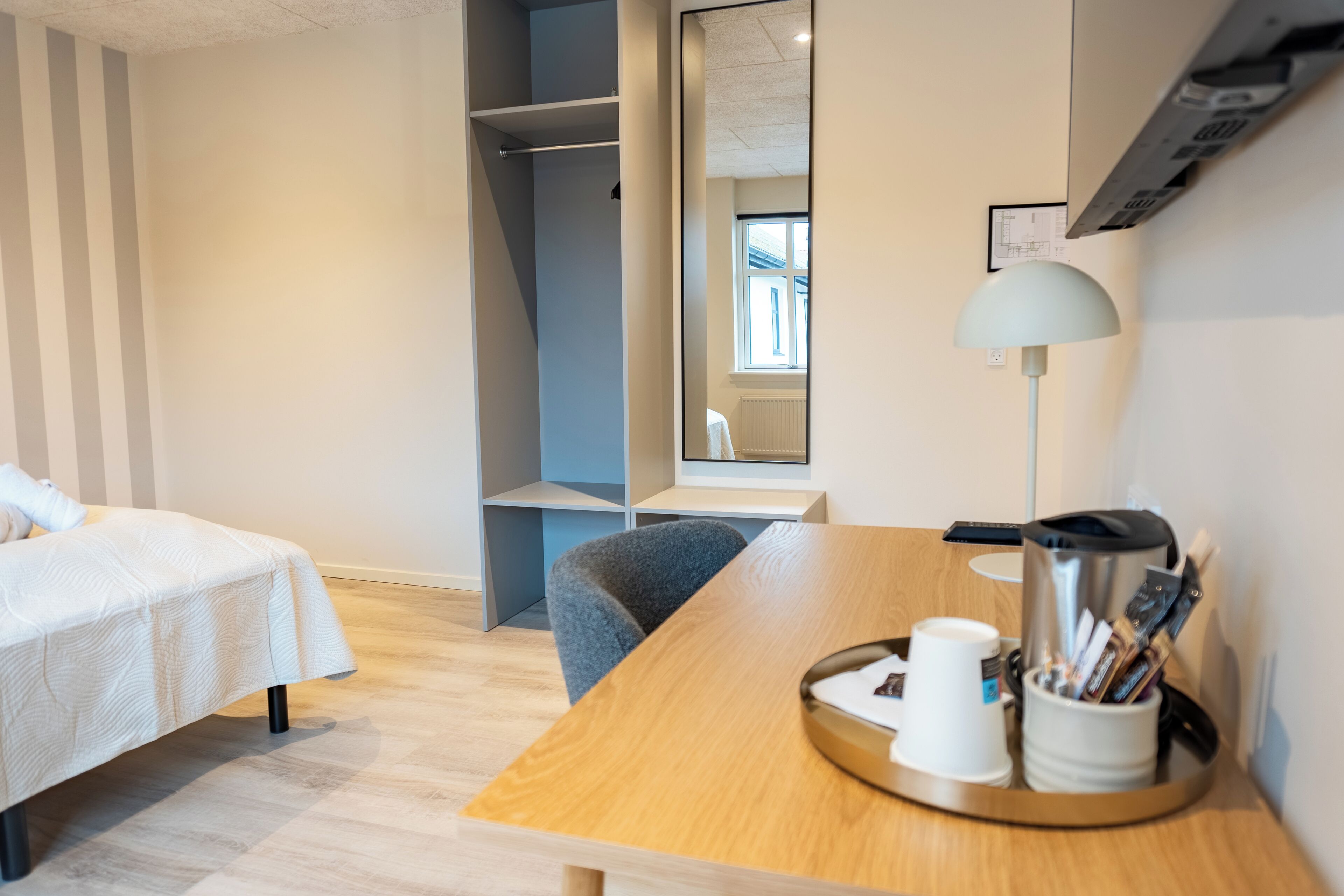 A hotel room with a bed, a desk with a lamp, a coffee maker, and a tray with cups and pens.