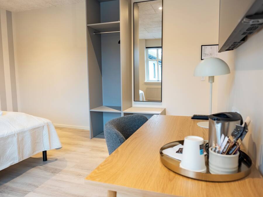A hotel room with a bed, a desk with a lamp, a coffee maker, and a tray with cups and pens.