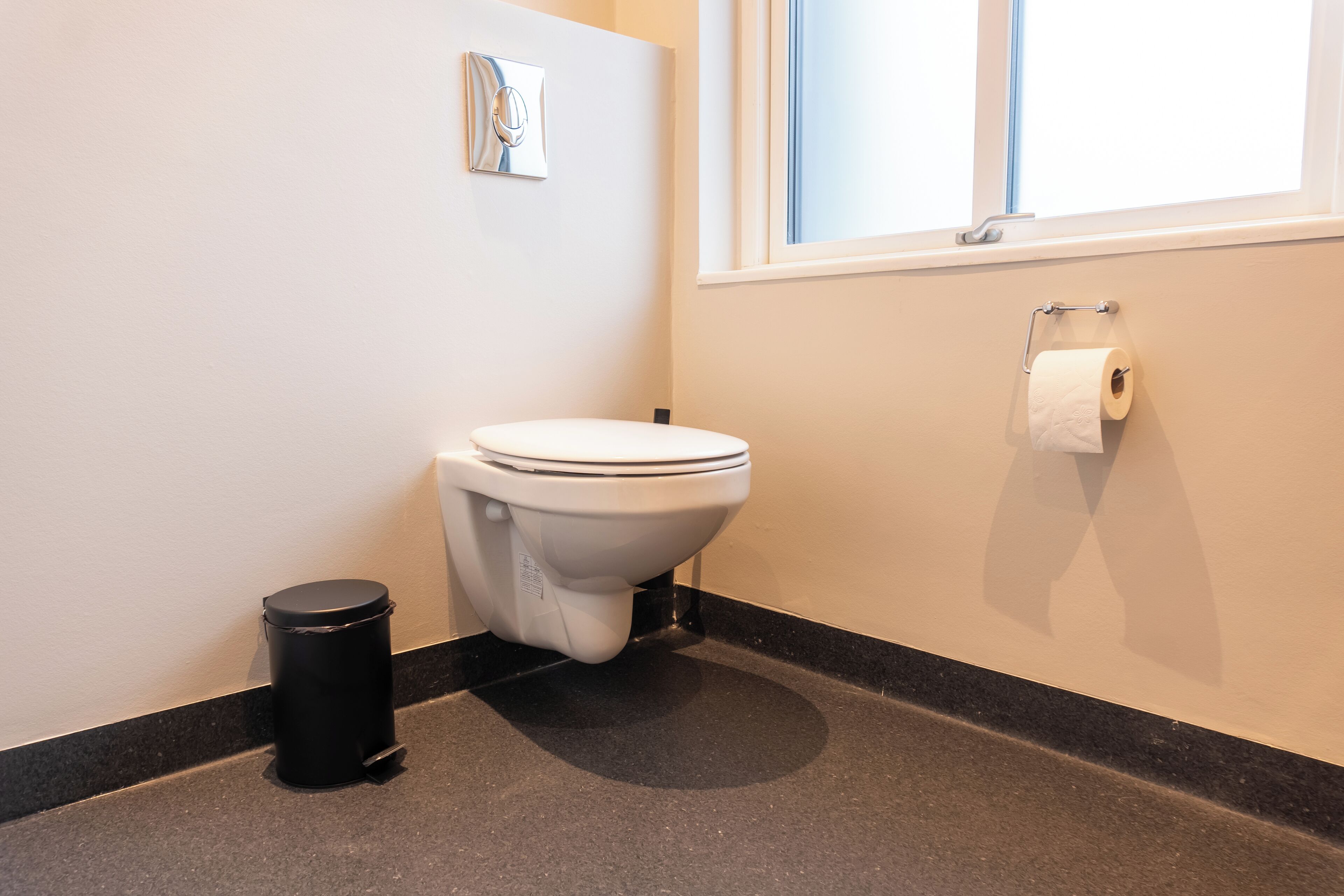 Une salle de bain équipée de toilettes, d’une poubelle, d’une fenêtre et d’un porte-rouleau de papier toilette.