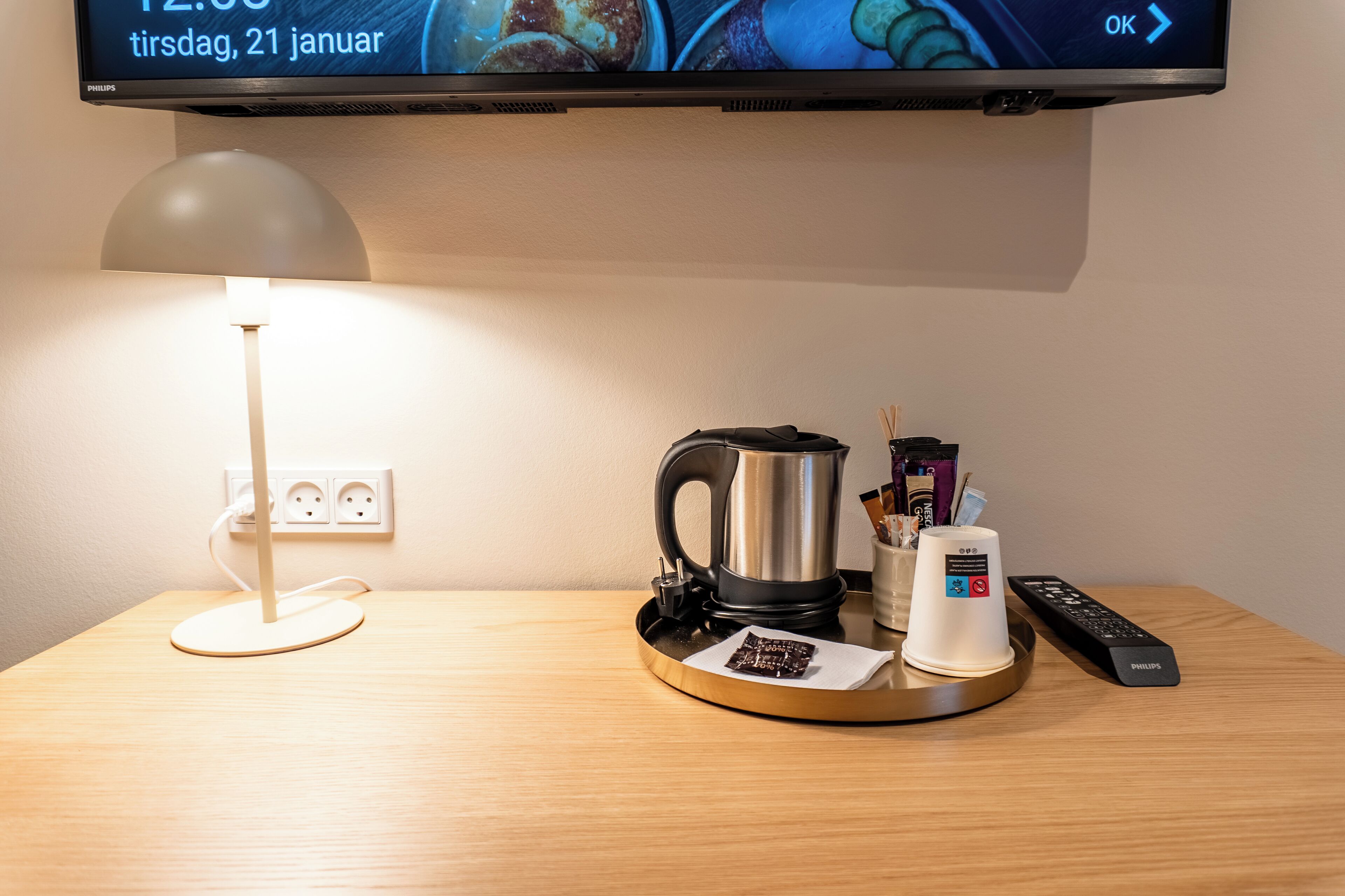 A hotel room with a television displaying a date, a coffee maker, a remote control, and a tray with a cup and a packet of snacks.