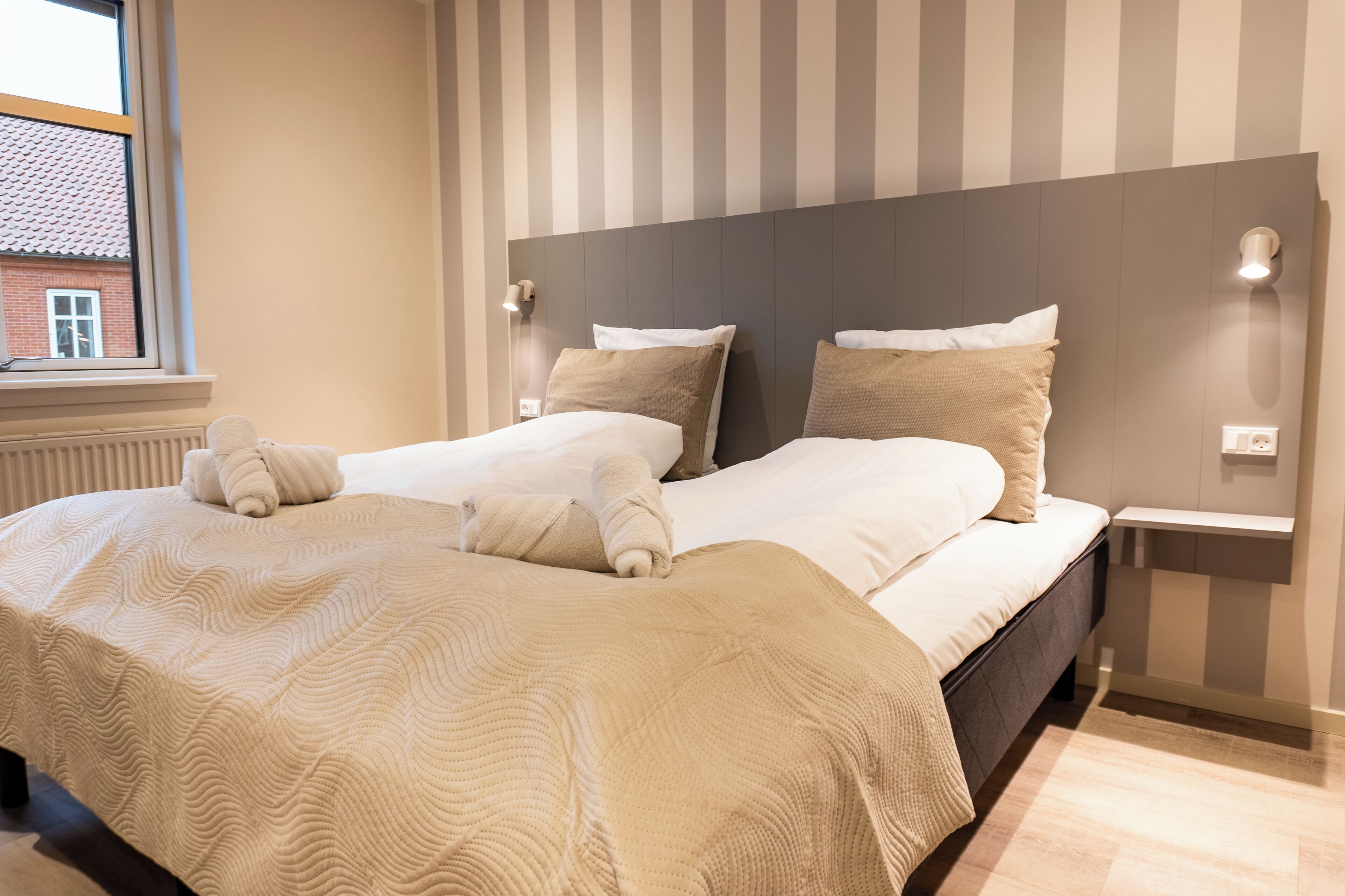 Een net opgemaakt bed met een gestreept hoofdbord, een nachtkastje met een lamp, en een raam met uitzicht op een gebouw.