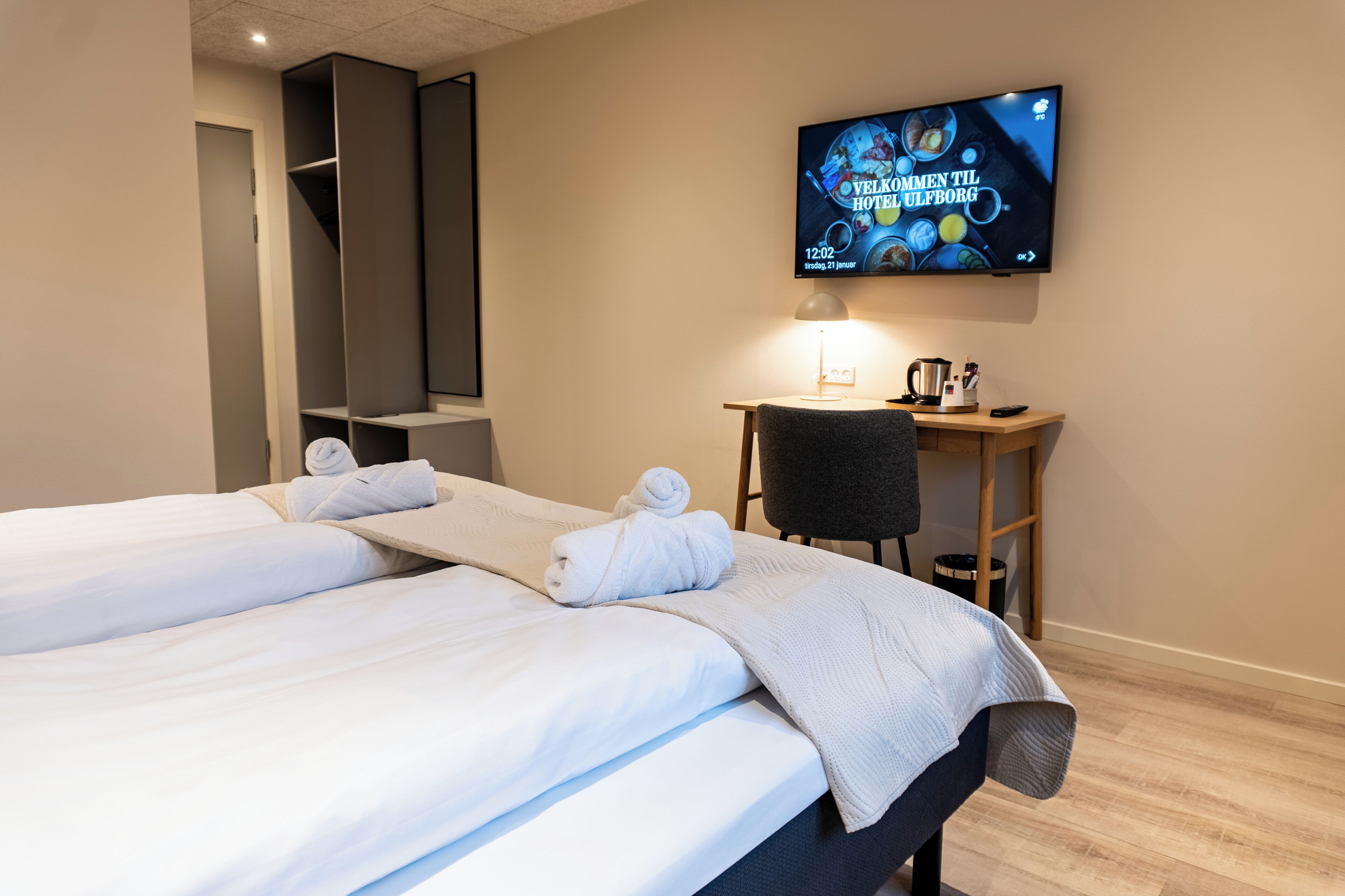 A hotel room with two beds, a TV mounted on the wall, a desk with a chair, and a small table with a kettle and cups.