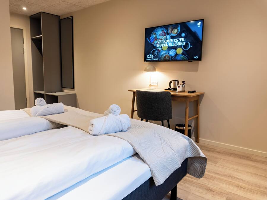 A hotel room with two beds, a TV mounted on the wall, a desk with a chair, and a small table with a kettle and cups.