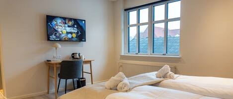A hotel room with two beds, a desk with a chair, a TV displaying a welcome message, and a window with a view of buildings.