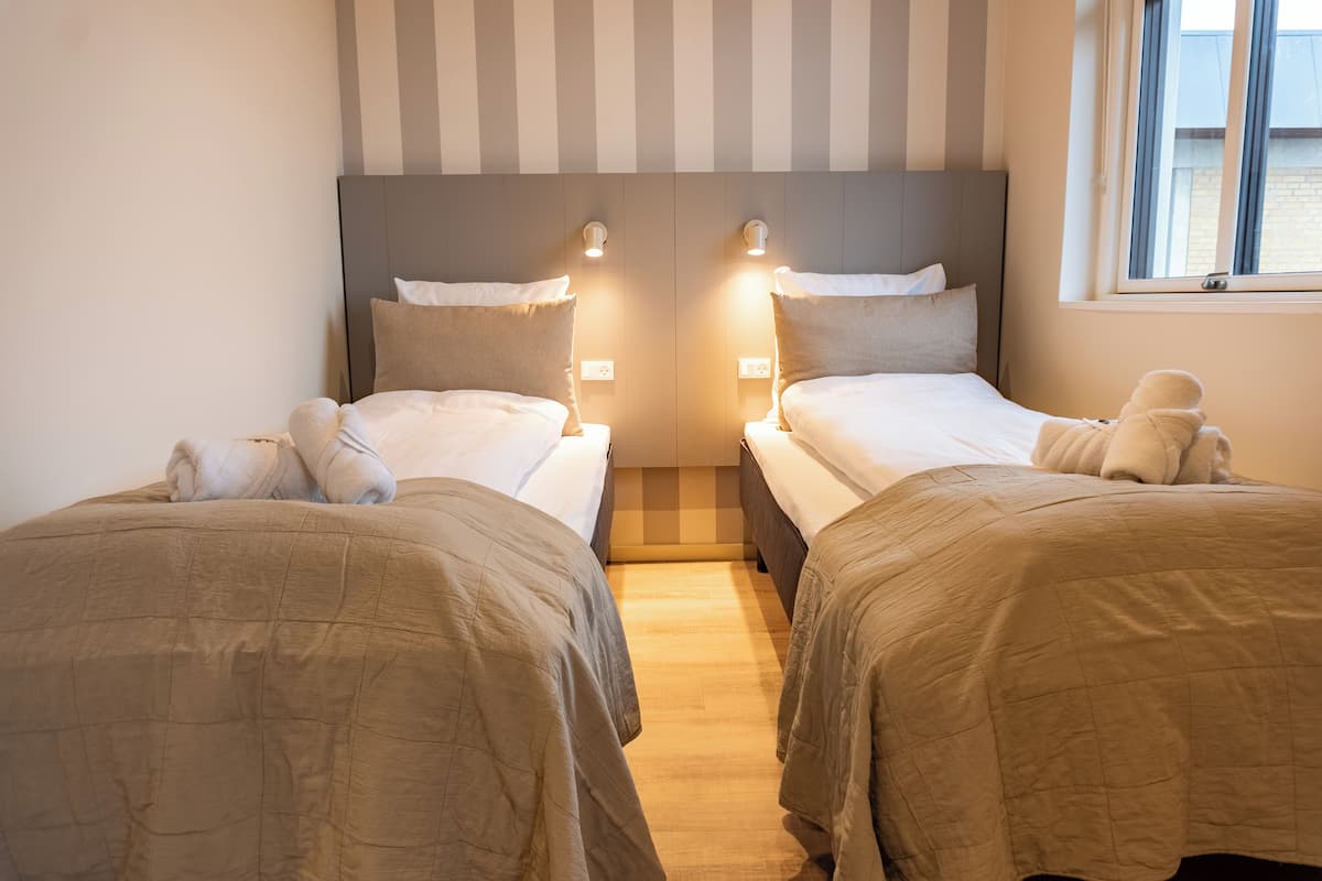 Ein Hotelzimmer mit zwei Betten, jeweils mit Kopfteil, Nachttischlampen und einem Fenster mit Vorhängen.
