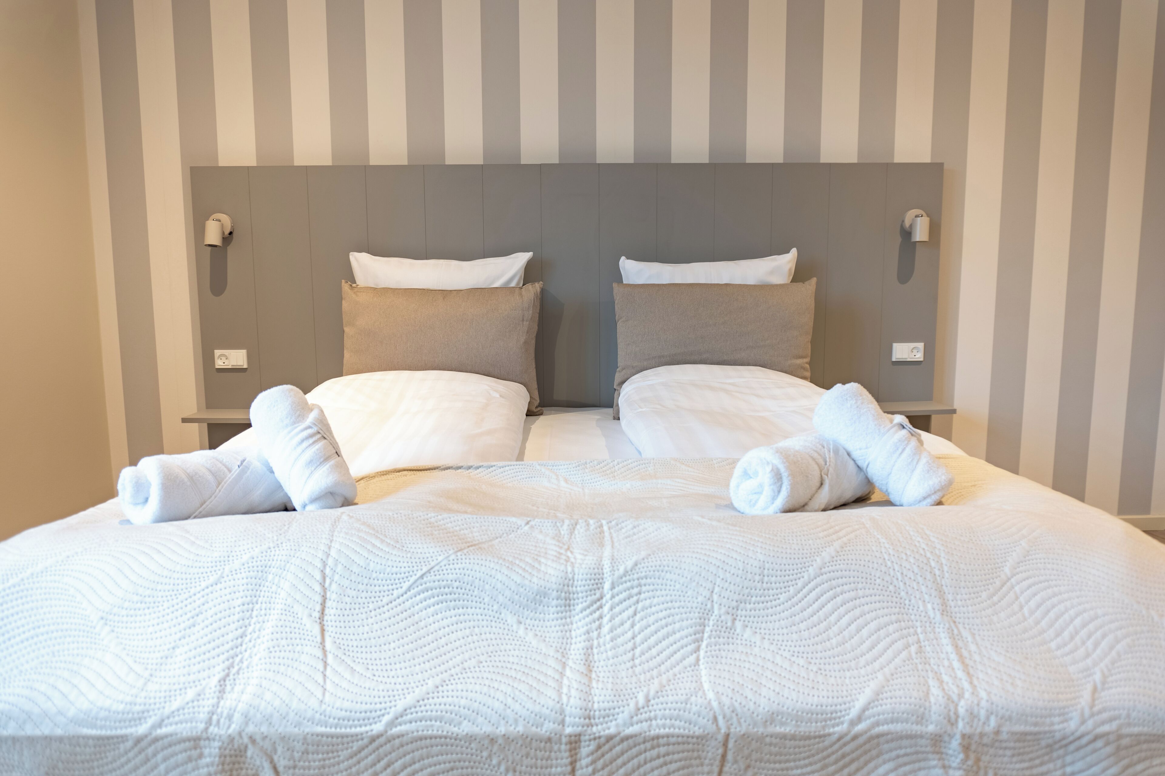 A double bed with white bedding and two pillows, a striped wall, and a headboard with light switches.