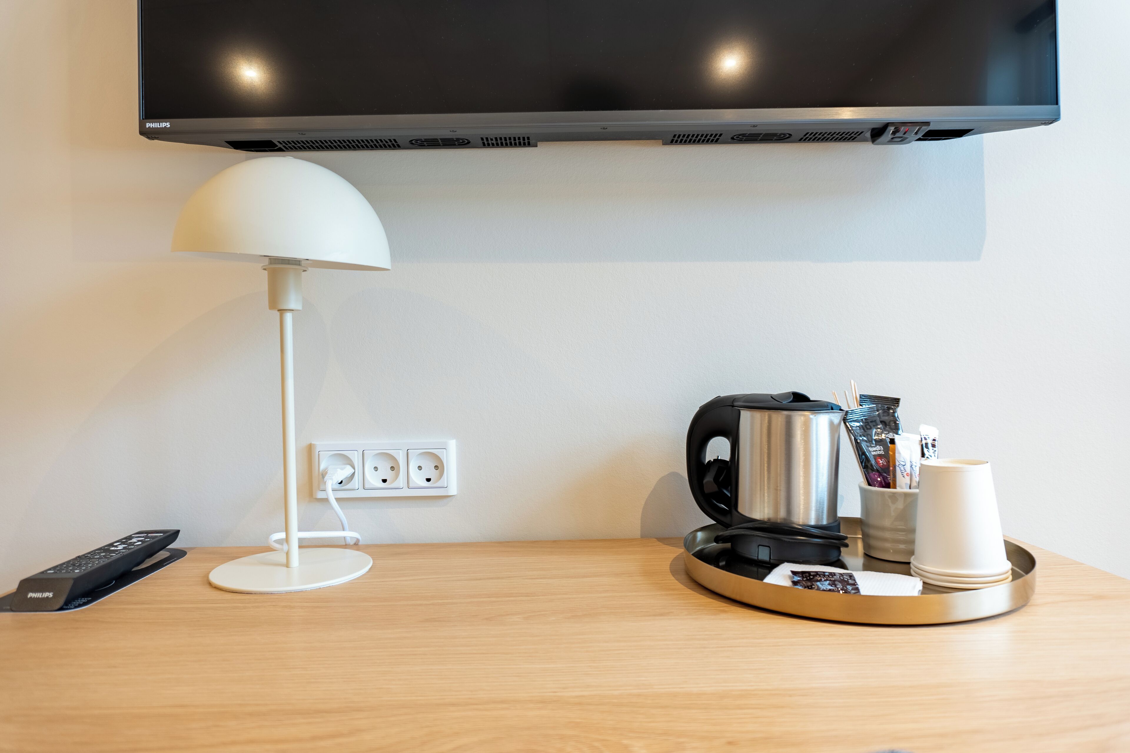 Camera d'albergo con televisione a schermo piatto montata a parete, una lampada bianca, una macchina del caffè, un vassoio con una tazza e un telecomando.