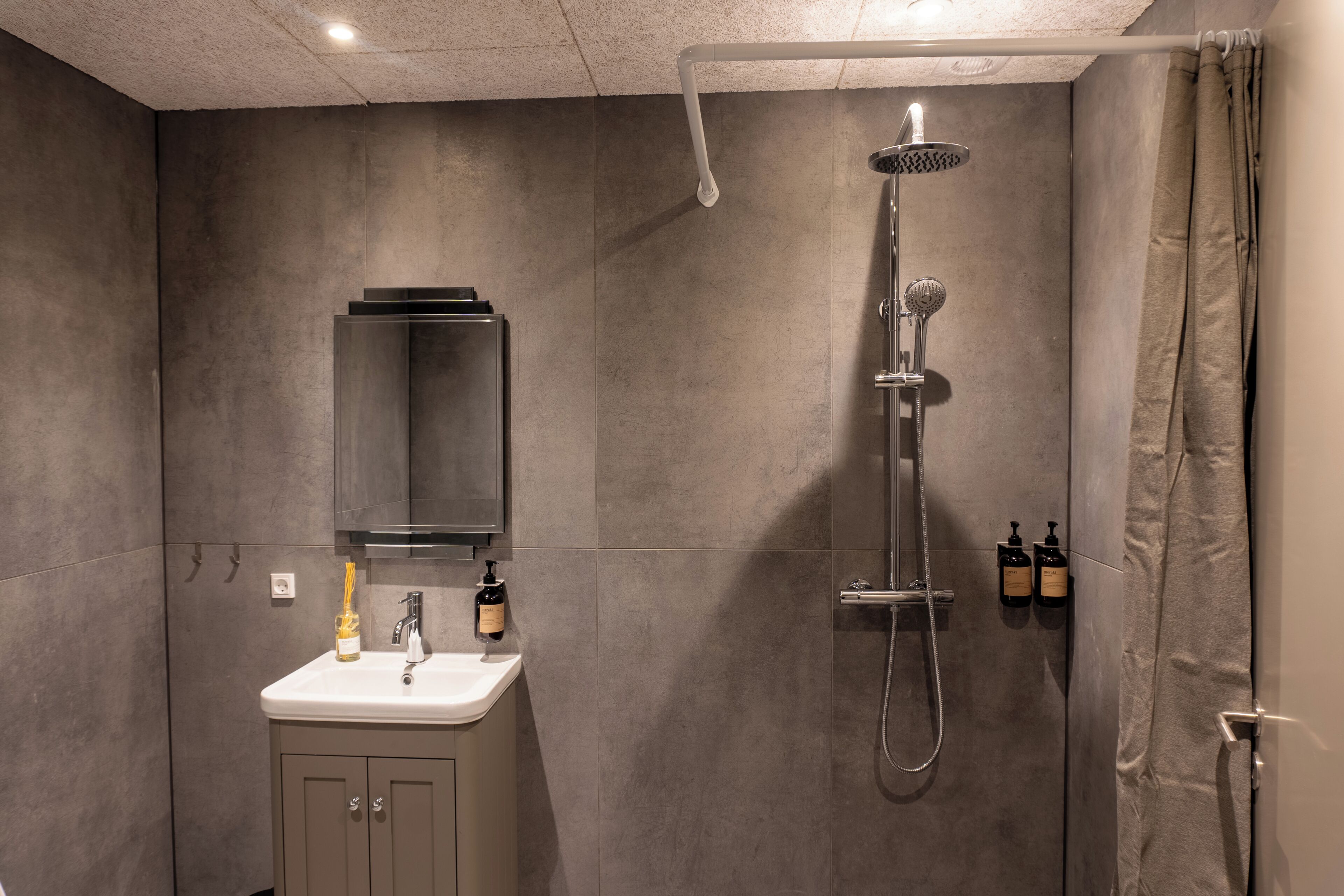 Un baño moderno con pared de azulejos grises, un lavabo blanco con espejo y una ducha con cabezal de ducha manual.