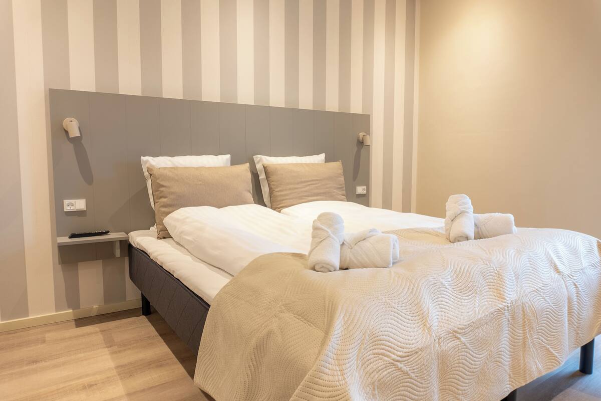 Ein ordentlich bezogenes Bett mit weißen Leinentüchern und zwei Kissen, ein Nachttisch mit Fernbedienung und eine Wand mit vertikalen Streifen.