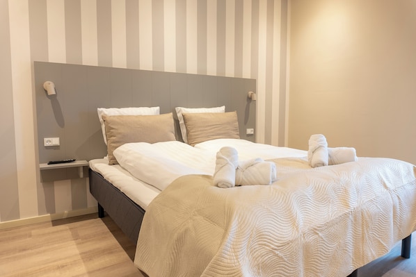 A neatly made bed with white linens and two pillows, a bedside table with a remote control, and a wall with vertical stripes. - Hotel Ulfborg (Ulfborg)