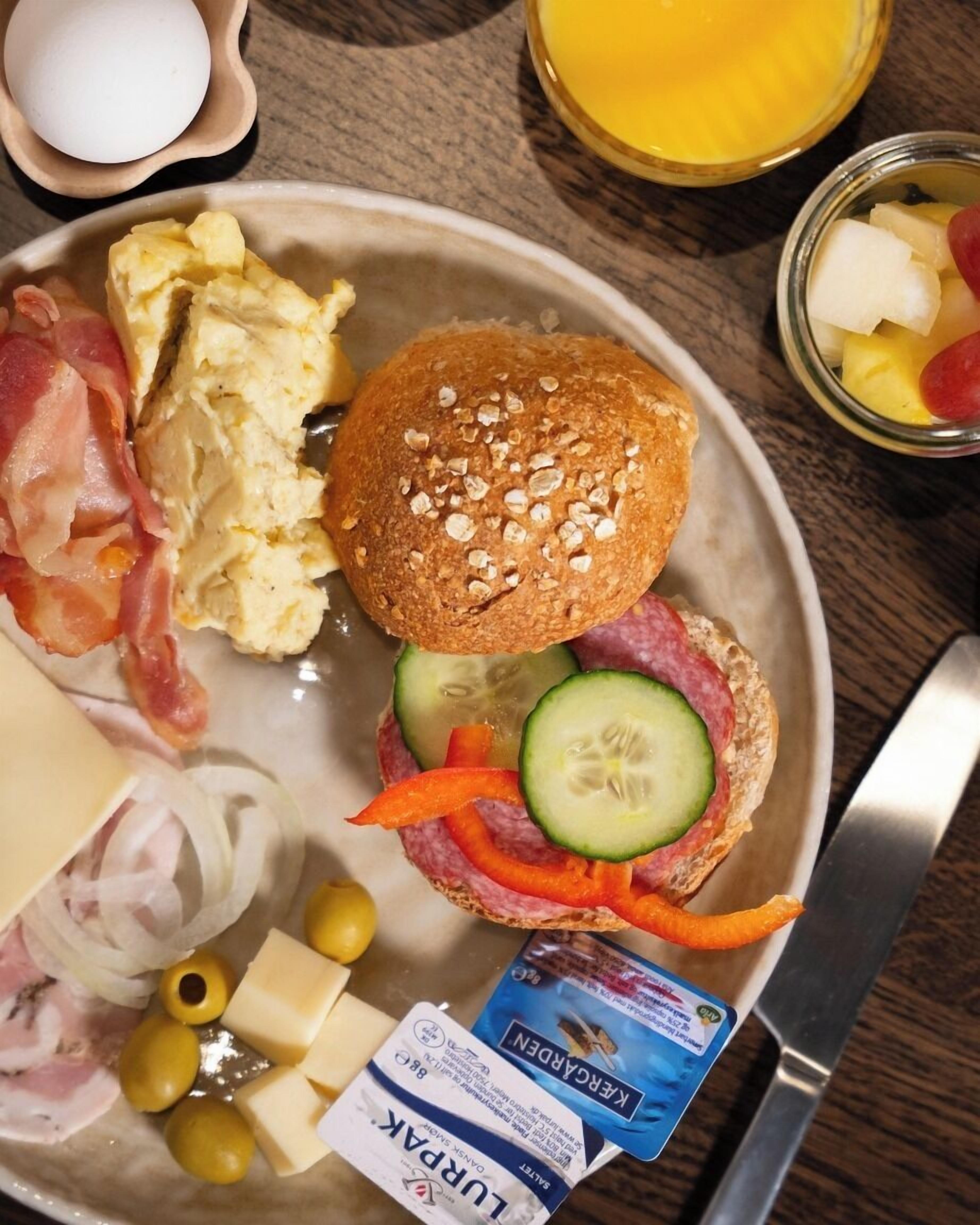 A breakfast plate with scrambled eggs, bacon, cheese, a sandwich with cucumber and tomato, and a glass of orange juice.