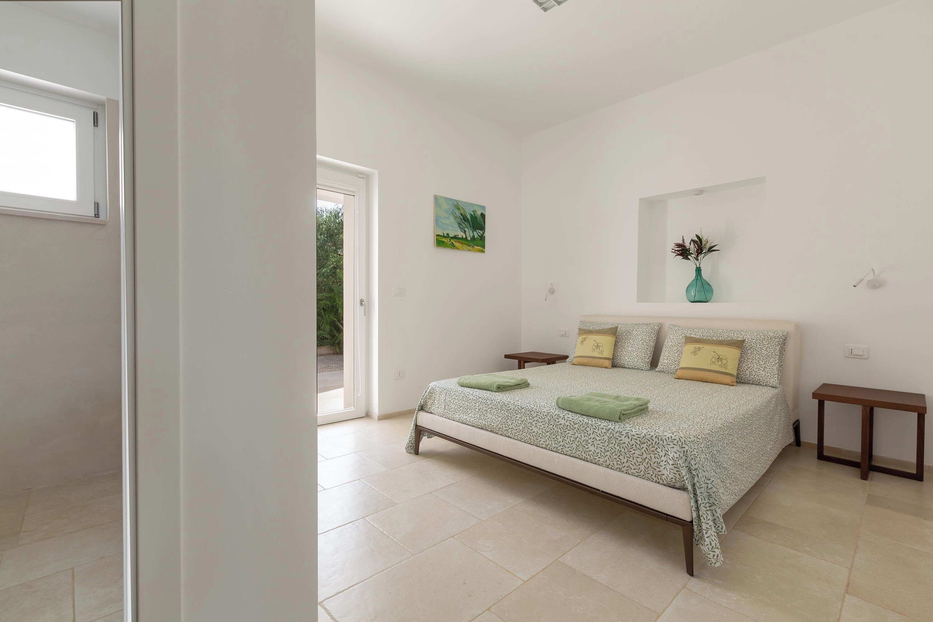 A bedroom with a bed, a nightstand, a small table, a painting, and a vase with flowers.