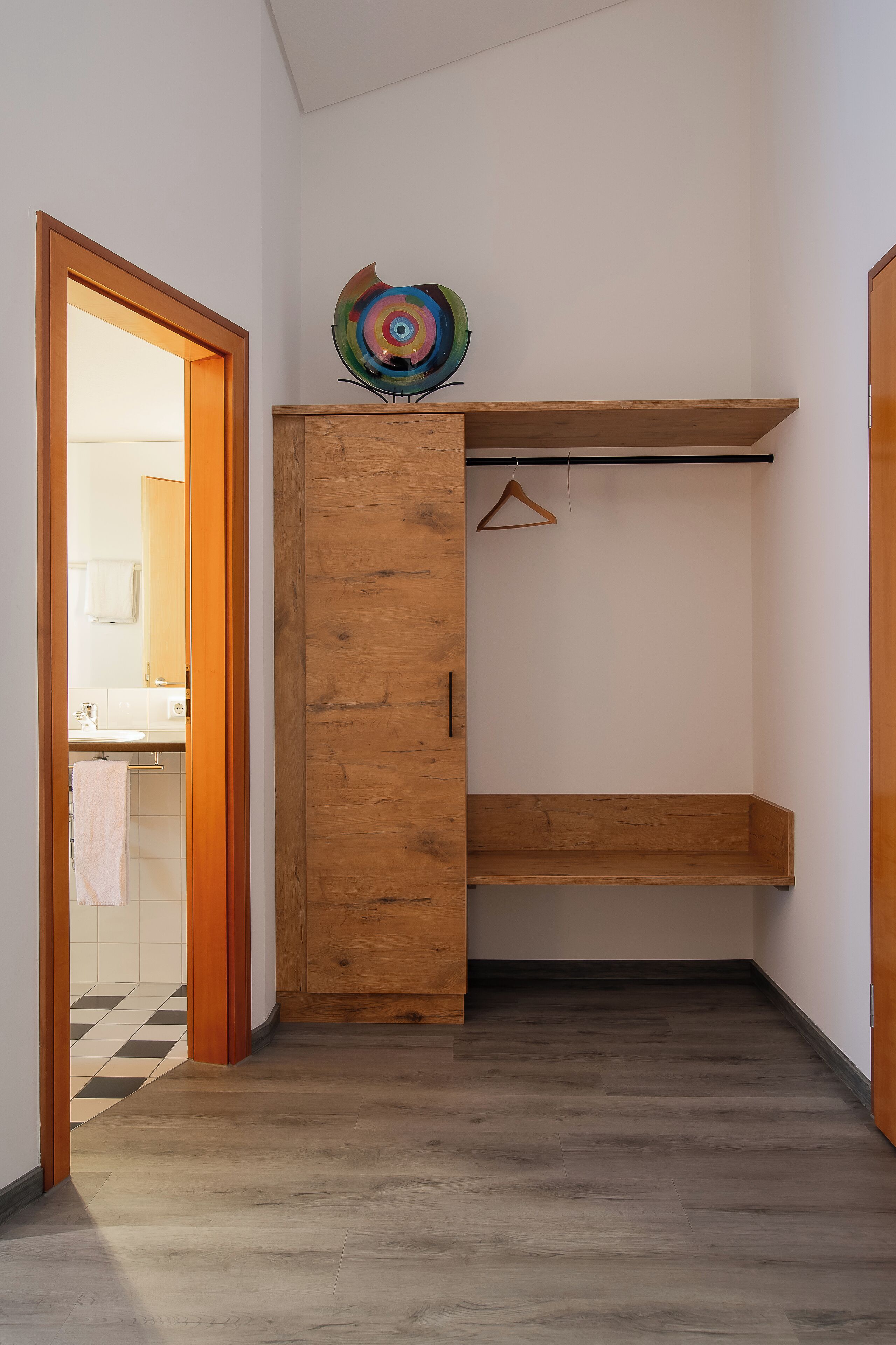 A hallway with a wooden wardrobe, a hanging coat rack, and a colorful abstract art piece on top.