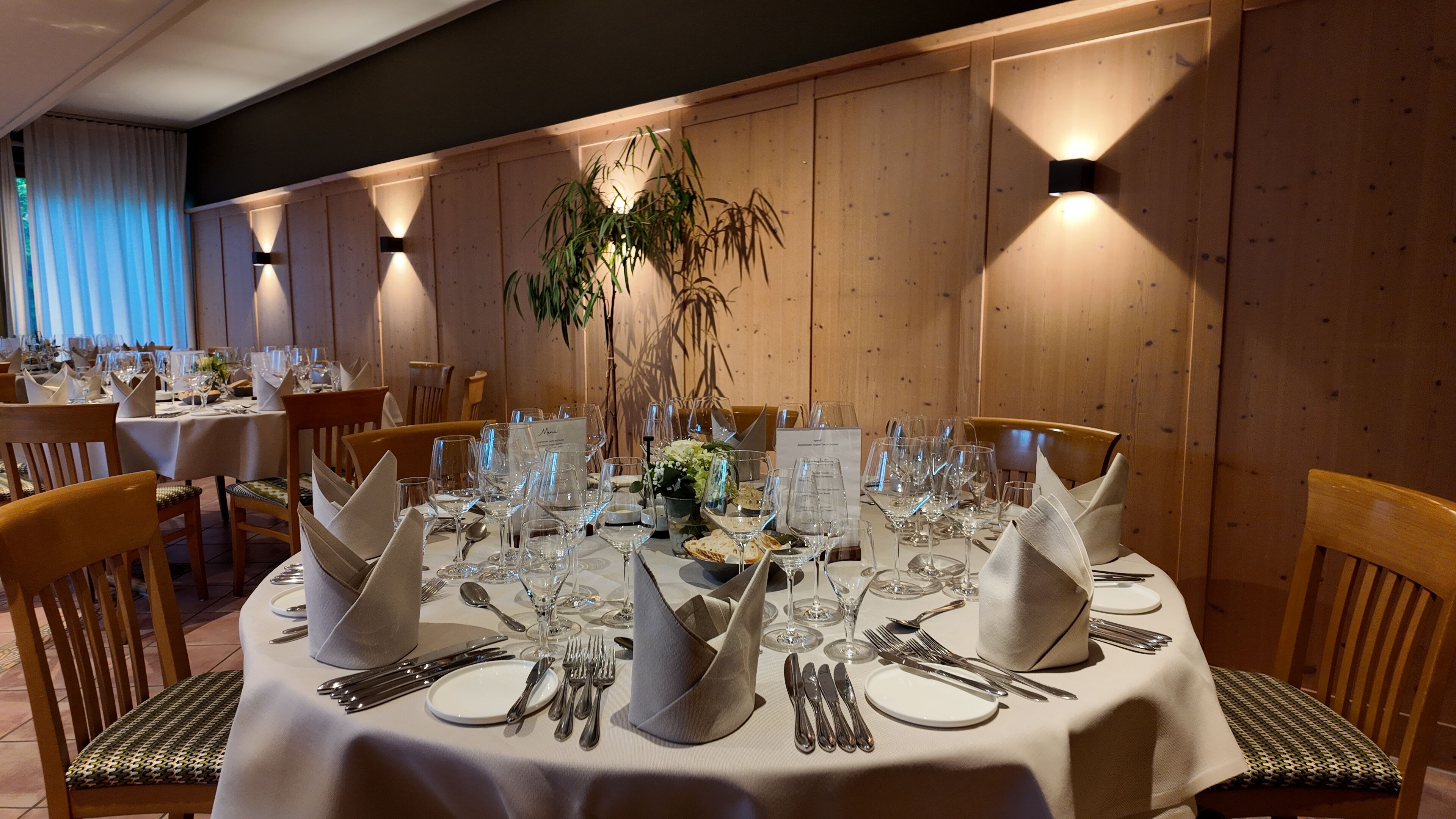 Comedor con mesas redondas dispuestas para una comida, con manteles blancos, cubertería ordenada y un arreglo floral central.