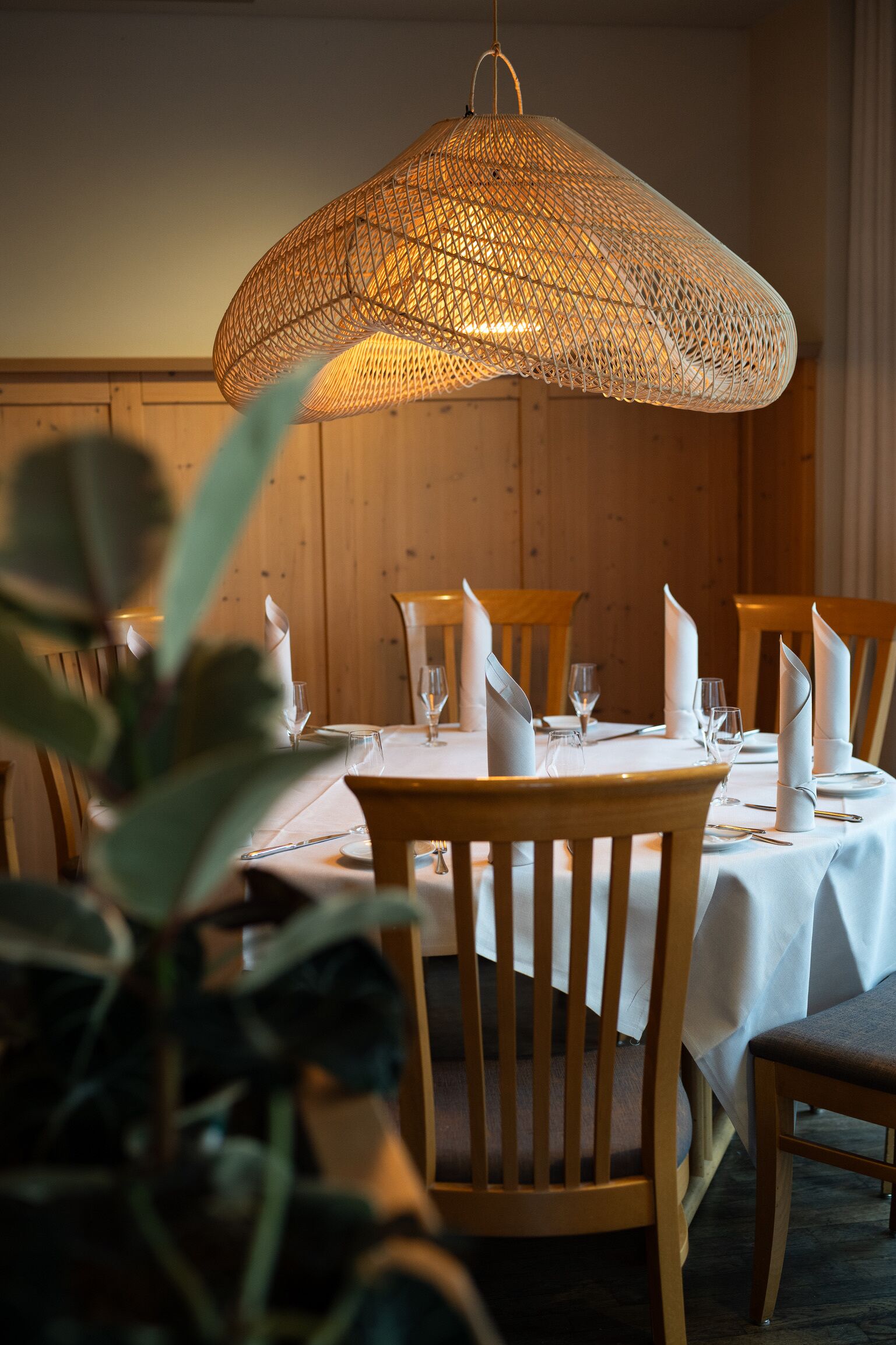 A dining area with wooden chairs, white tablecloths, and a woven pendant light.