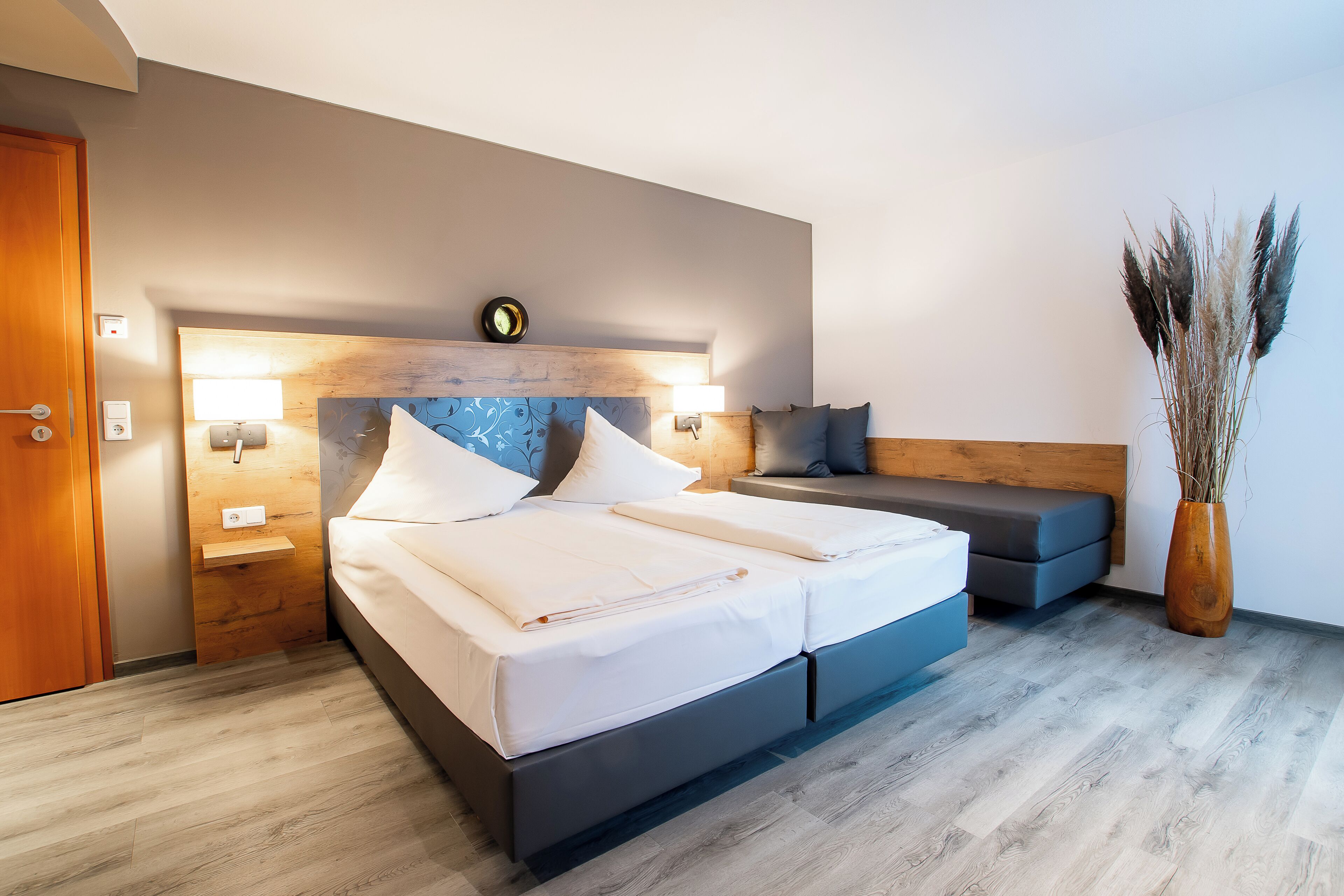 Un dormitorio moderno con cama, mesitas de noche, una banqueta y un florero con plantas secas.