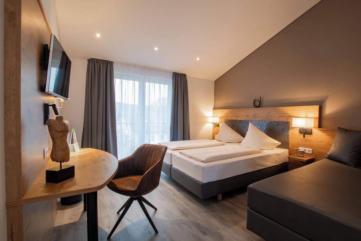 A hotel room with a bed, a desk with a chair, a TV mounted on the wall, and a window with curtains.