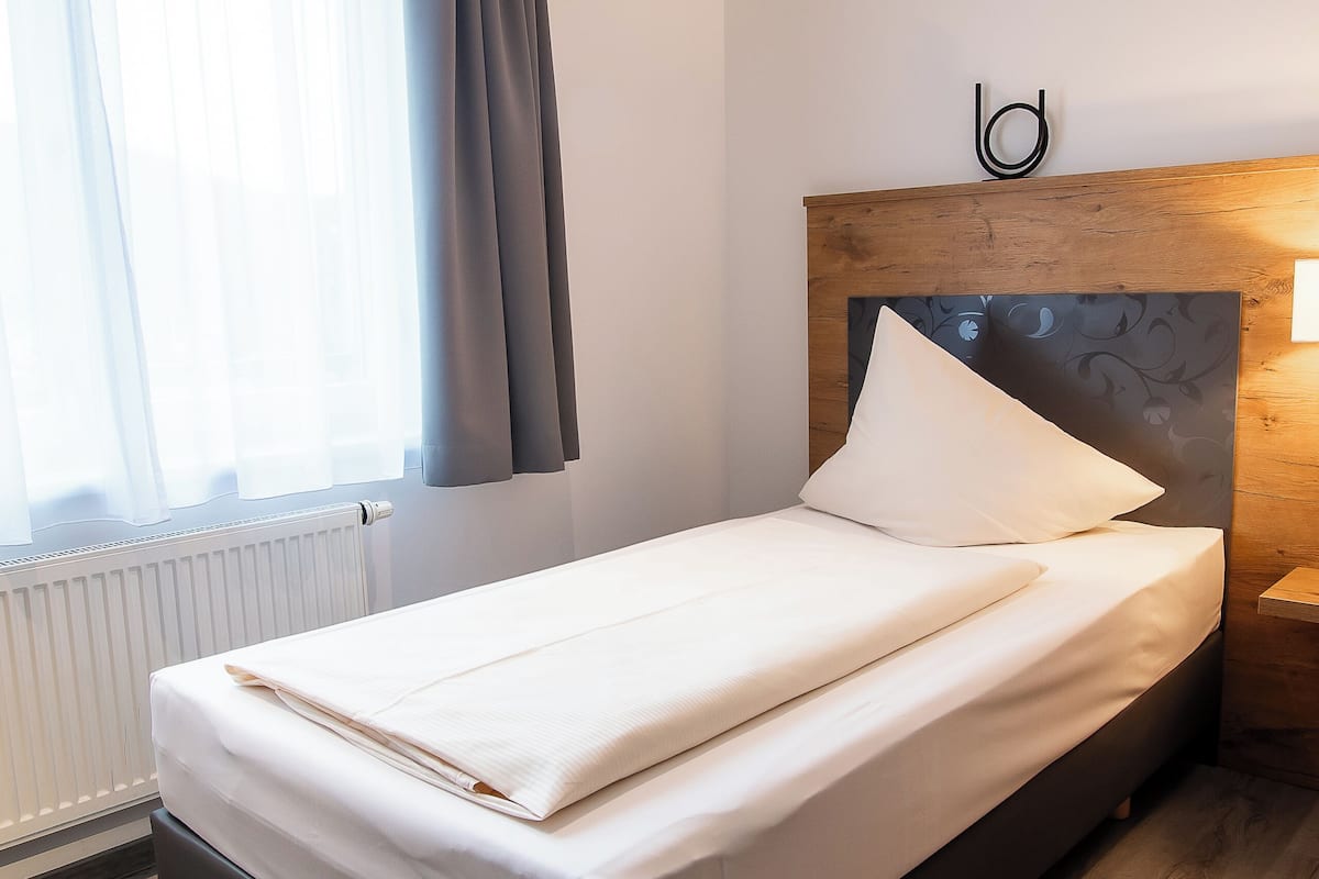 A neatly made bed with a white comforter and pillows, a wooden headboard with a letter 'b', a bedside table with a lamp, and a window with curtains.
