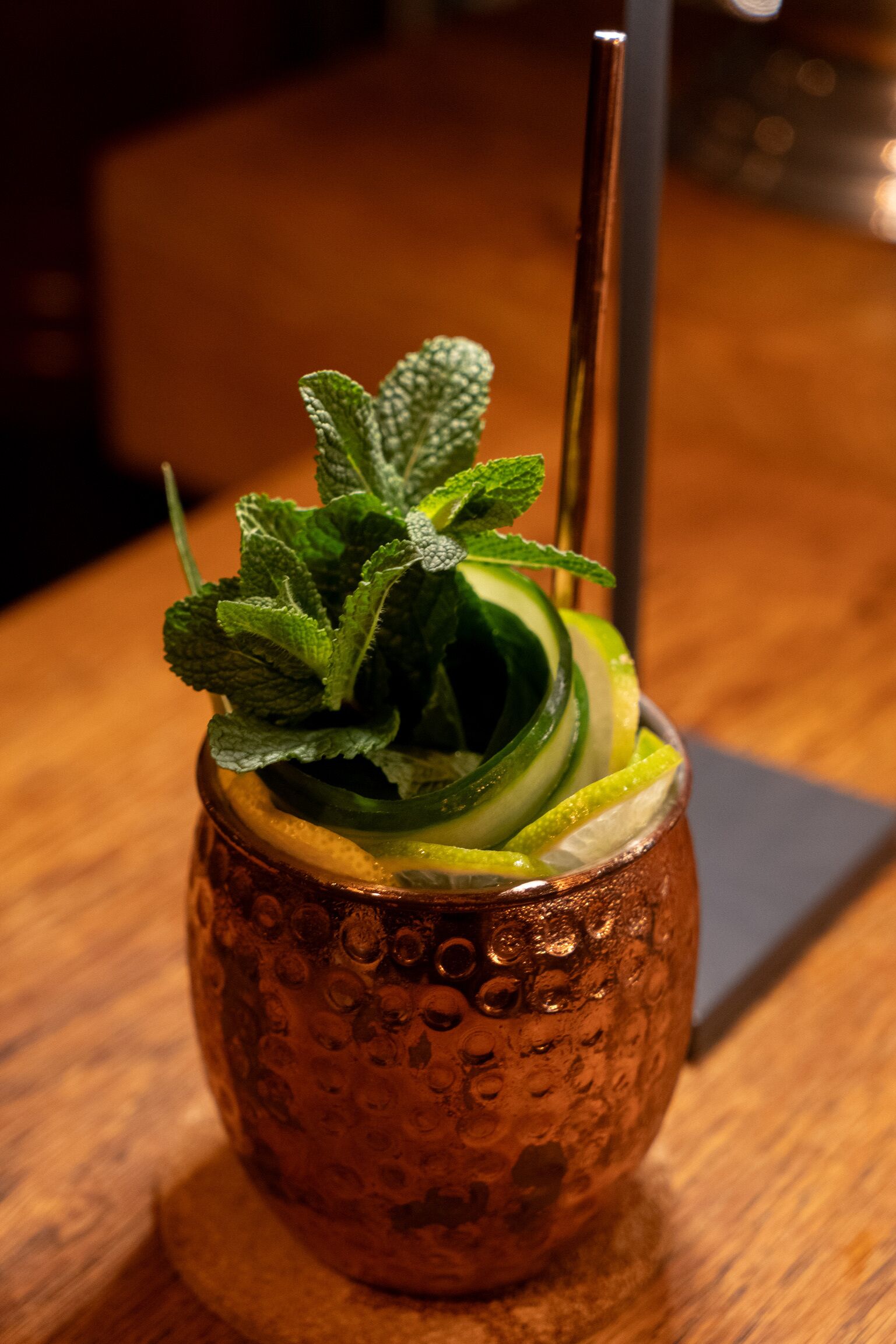 A copper mug with a textured pattern, containing a beverage garnished with mint leaves, cucumber slices, and a lemon wedge, with a straw.