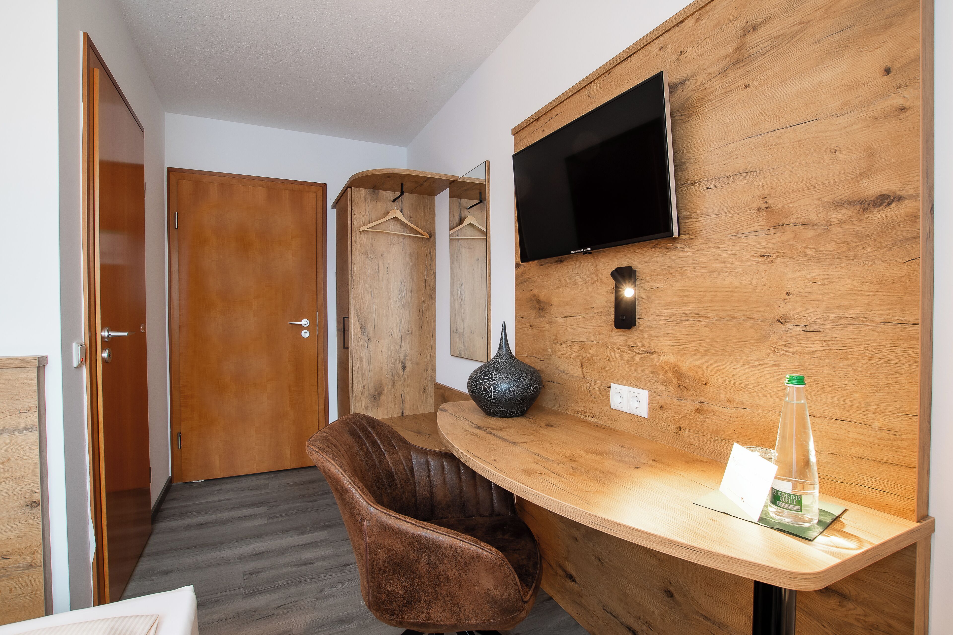 A hotel room with a wooden desk, a brown leather chair, a flat-screen TV mounted on the wall, and a wooden wardrobe.