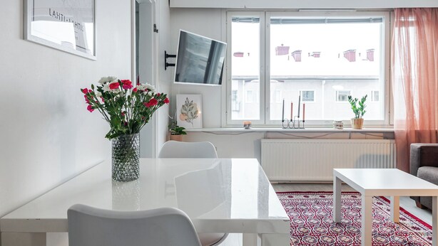 Una sala de estar moderna con una mesa blanca, sillas, un televisor montado en la pared y un ventanal con vista a edificios.