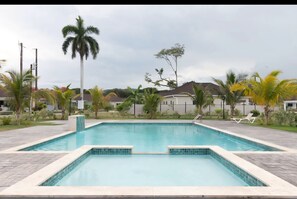 Une piscine rectangulaire dont les bords sont recouverts de carreaux, entourée d’une zone pavée agrémentée de palmiers et d’une clôture.